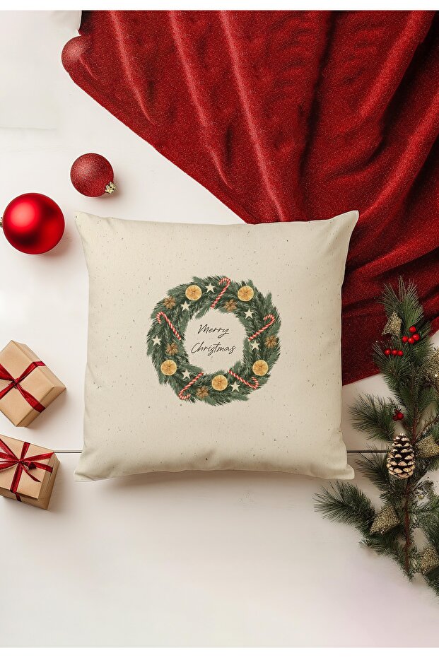 Decorative pillow with Christmas wreath design on a white surface with red blanket and ornaments.
