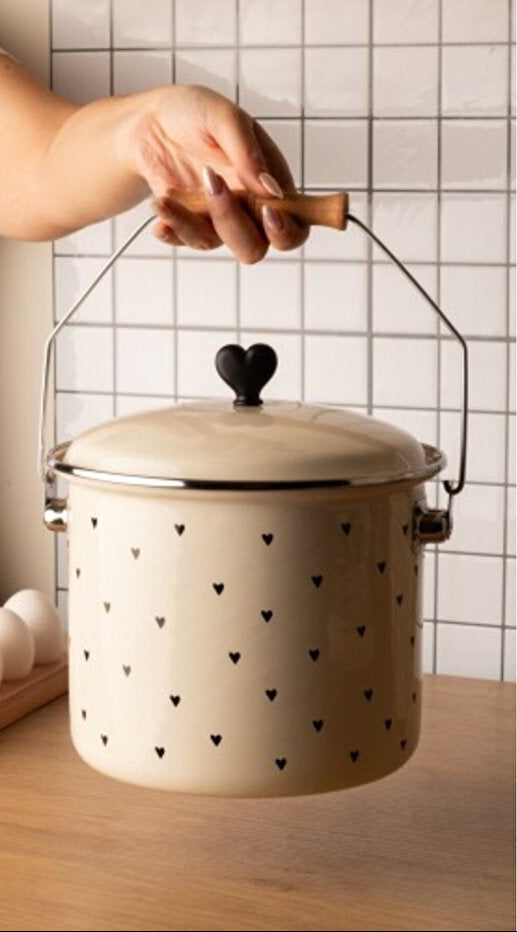 Beige enamel pot with black heart patterns and a wooden handle on a tiled wall background.