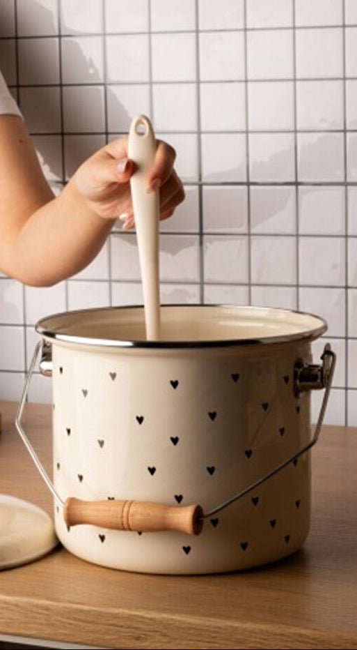Person using a wooden spoon to stir contents in a beige pot with black heart patterns on a tiled wall background.